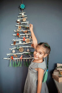 Portrait of smiling girl holding gift against christmas tree hanging on wall