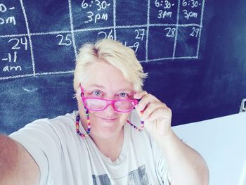 High angle portrait of smiling teacher wearing eyeglasses against blackboard