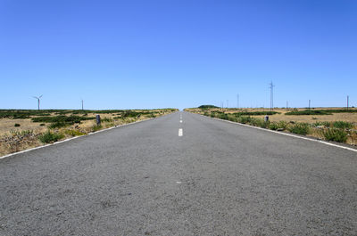 Empty road amidst land against clear blue sky