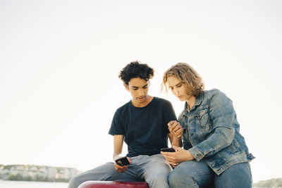 Friends using mobile phone while sitting on bollard against clear sky in city