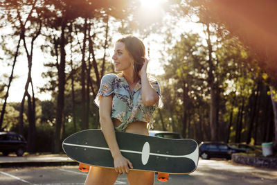 Cheerful woman looking away outdoors while holding black skateboard