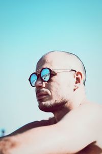 Side view of shirtless mid adult man wearing sunglasses while sitting at beach against clear sky during sunny day