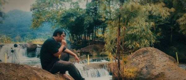 Side view of young woman sitting on rock against trees