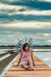 Portrait of mid adult woman sitting on pier over lake against cloudy sky during sunset