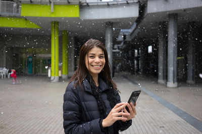 Portrait of smiling young woman using mobile phone in city during winter