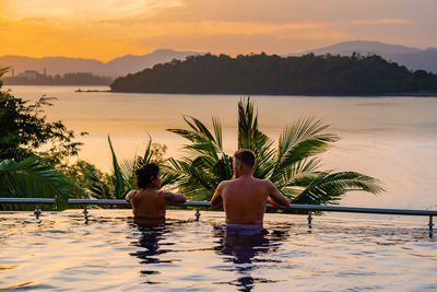 Rear view of shirtless man sitting in swimming pool against sunset sky