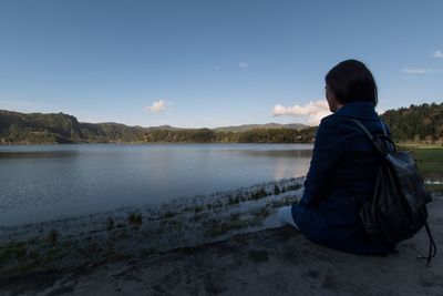 Rear view of woman sitting by lake against sky