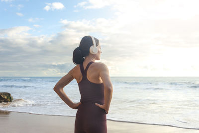 Fit, sporty woman standing by the ocean with hands on her hips and wearing headphones
