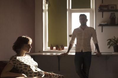 Pregnant woman sitting while man standing in background at home