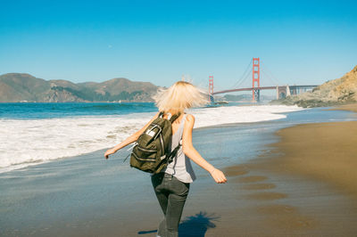 Rear view of woman walking on shore with golden gate bridge against clear sky