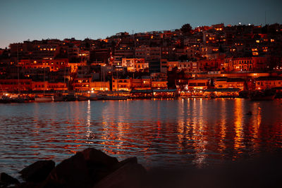 Illuminated buildings by sea against sky during sunset