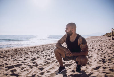 Young man sitting on sand at beach against clear sky