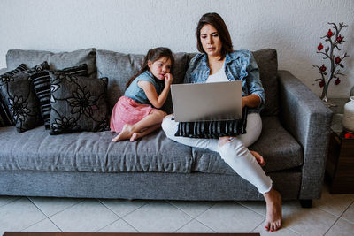 Full body of young latin american mother in casual clothes sitting on sofa and watching interesting video with cute little daughter