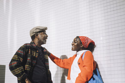 Happy father and daughter talking with each other while standing against wall during sunny day