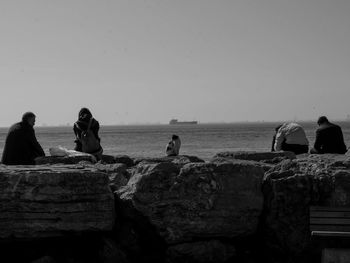 People sitting on rock by sea against clear sky