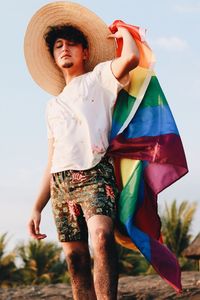 Young man holding rainbow flag while standing at beach