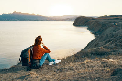 Man sitting on shore at beach against sky during sunset