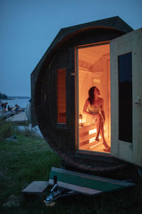 Full length of smiling woman sitting in sauna cabin by sea against sky