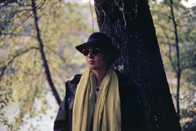 Woman wearing sunglasses standing against tree trunk in forest