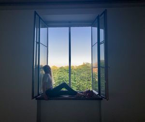 Woman sitting on window glass against clear sky