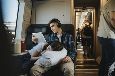 Girl resting on brother's lap using tablet pc while sitting on seat in train