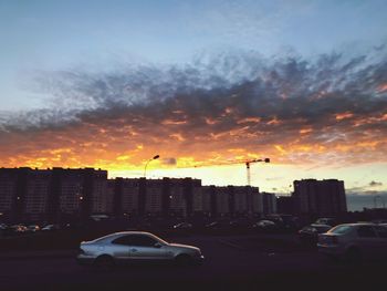 Cars on road by buildings against sky during sunset