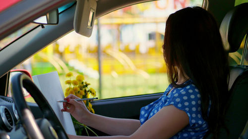 Beautiful brunette woman, sits in car, draws pencil sketch, drawing bouquet of yellow dandelions.