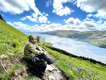 Young man sitting on rock against sky