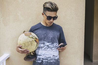 Teenage boy using mobile phone while holding globe against wall