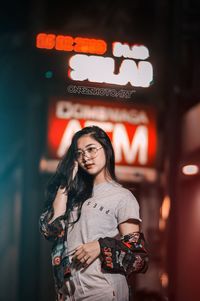 Portrait of young woman standing against illuminated wall at night