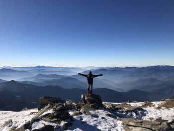 Rear view of man with arms outstretched looking at mountain ranges against clear blue sky