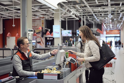 Mature cashier talking to woman while paying at checkout counter in supermarket