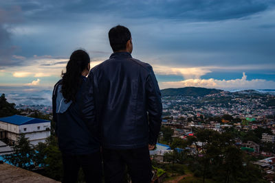 Young couple watching downtown city view with dramatic cloudy sky at evening from mountain top