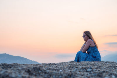 Side view of woman sitting on rock against sky during sunset