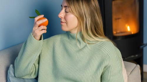 Blonde inhales the aroma of tangerine sitting on a sofa by the fireplace.