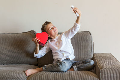 Boy holding heart shape doing selfie while sitting on sofa at home