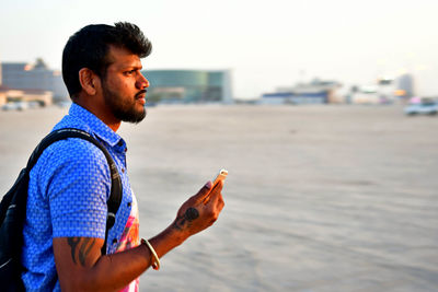 Side view of young man holding mobile phone while standing by river in city