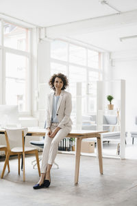 Businesswoman in office sitting on desk, looking confident