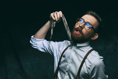 Young man holding necktie while making face against black background