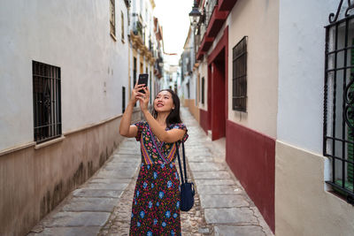 Cheerful asian female tourist taking picture of aged buildings while standing on paved narrow street of cordoba town in spain