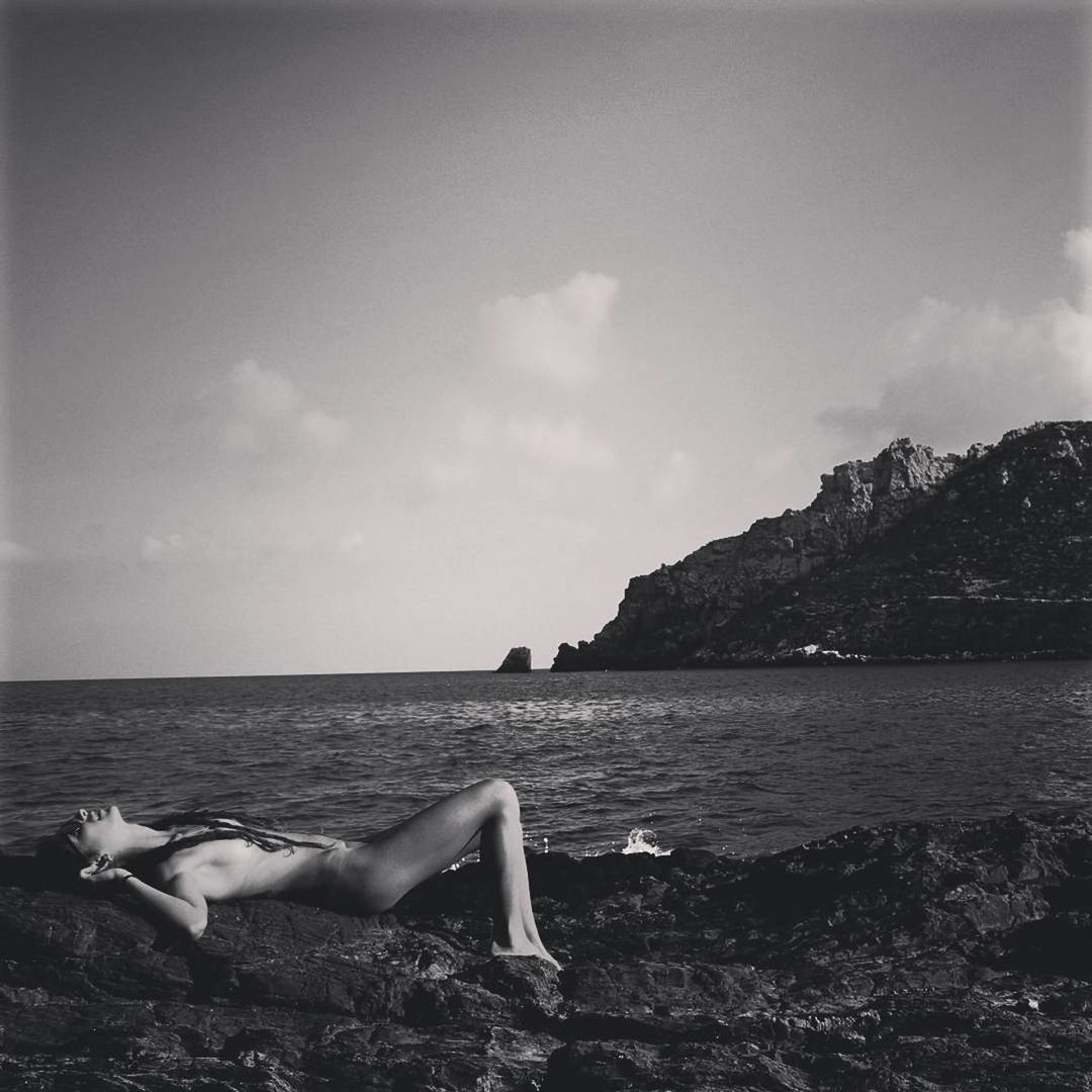 YOUNG WOMAN RELAXING ON ROCK BY SEA AGAINST SKY