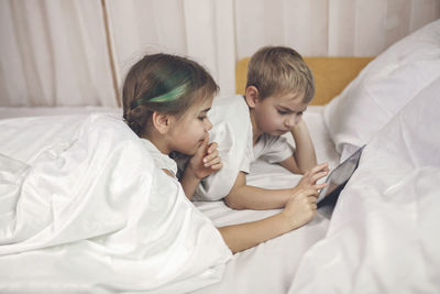 Cute sibling using digital tablet while lying on bed at home