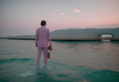 Rear view of man standing in sea against sky during sunset