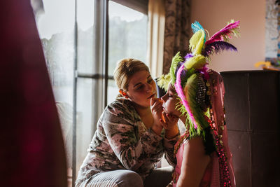 Side view of adult woman applying makeup on face of girl in colorful costume during festival preparation at home