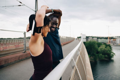 Smiling male and female athletes stretching hands on footbridge over sea against sky