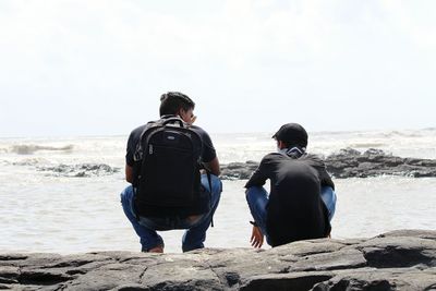 Rear view of friends crouching on rock by sea against clear sky