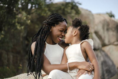 Happy black mother with afro braids smiling and kissing cheerful daughter in white clothes while spending summer weekend day in countryside
