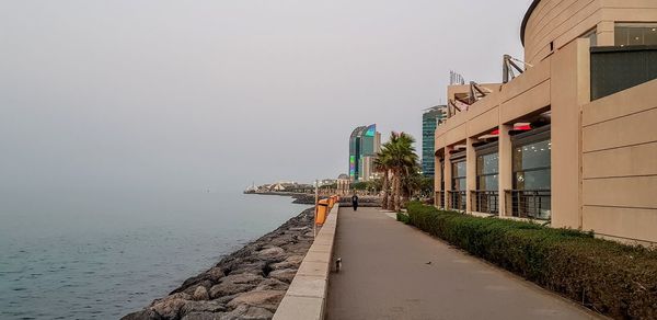 Footpath by sea against buildings in city against clear sky