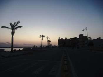 Street amidst silhouette trees against clear sky at sunset