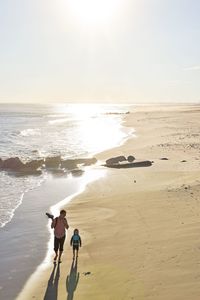 Man walking with son on shore at beach against sky on sunny day